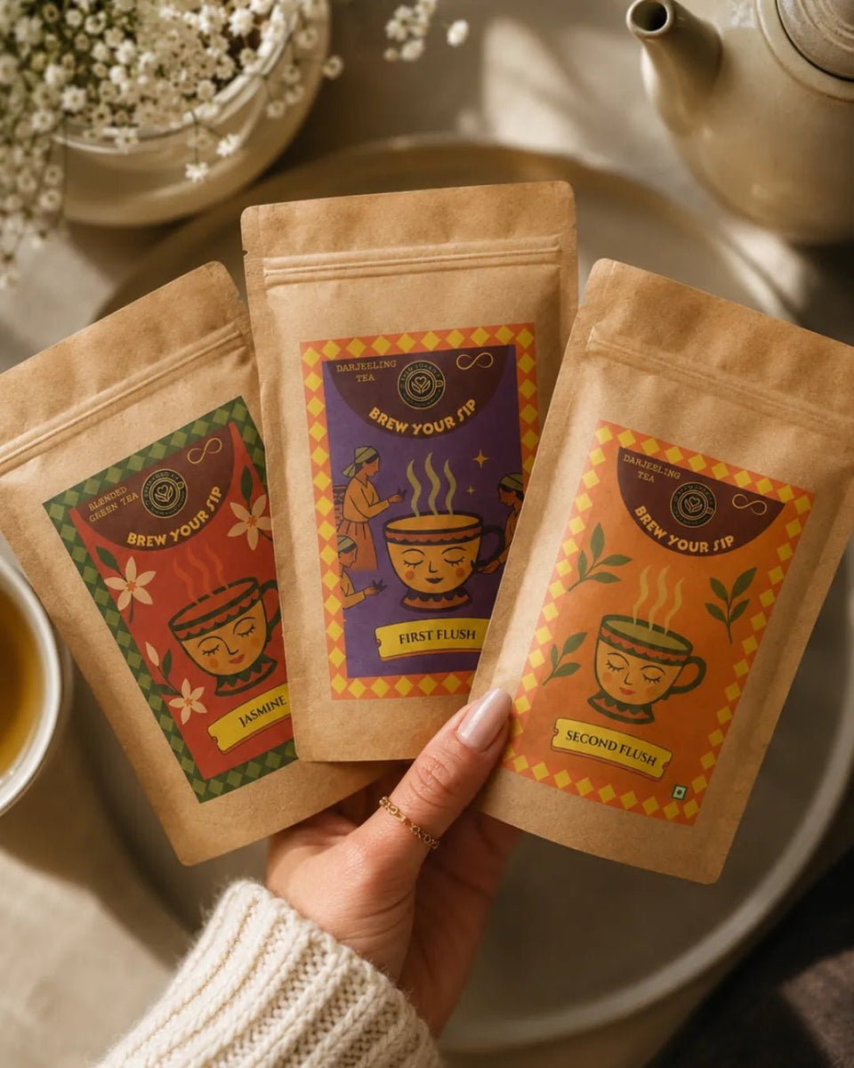 Hands holding three Brew Your Sip Darjeeling sampler pouches above a table set for tea